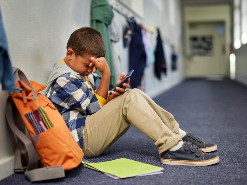 Little,Boy,With,Backpack,Holding,Smartphone,Sitting,Alone,In,School